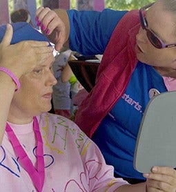 A middle aged woman at an event checks her forehead in the mirror while being assisted by another woman.